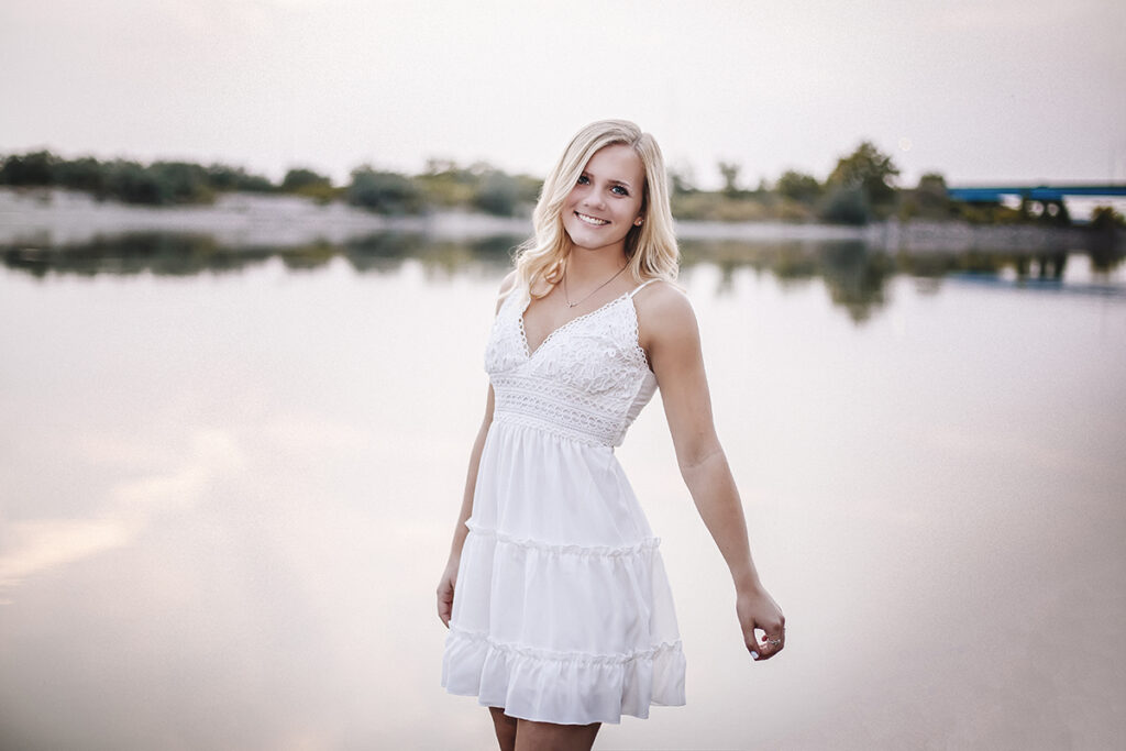 Young lady by river in white dress, smiling at camera.